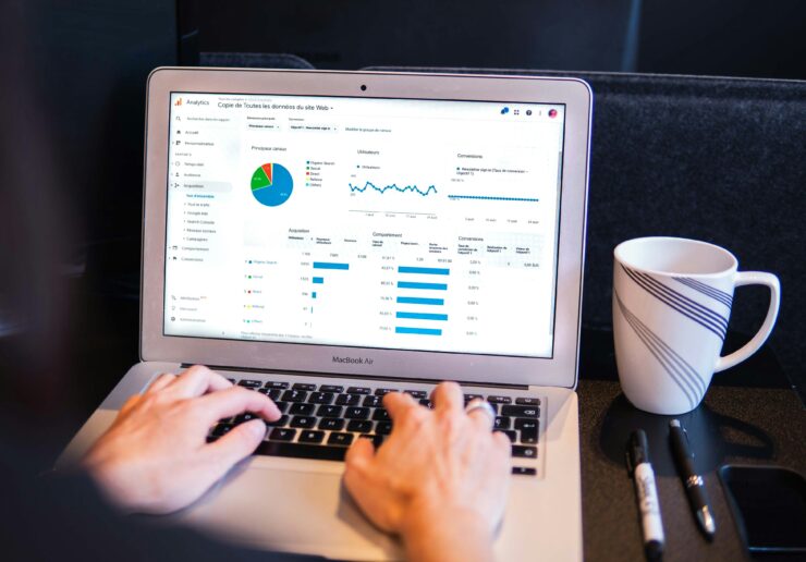 2 hands typing on a laptop keyboard with data analytics on screen and a mug and pens to the side